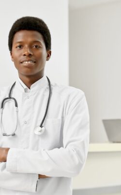 Selective focus of professional therapist with stethoscope on neck posing with arms crossed and looking at camera. Portrait of smiling african male doctor in white lab coat, reception on background.